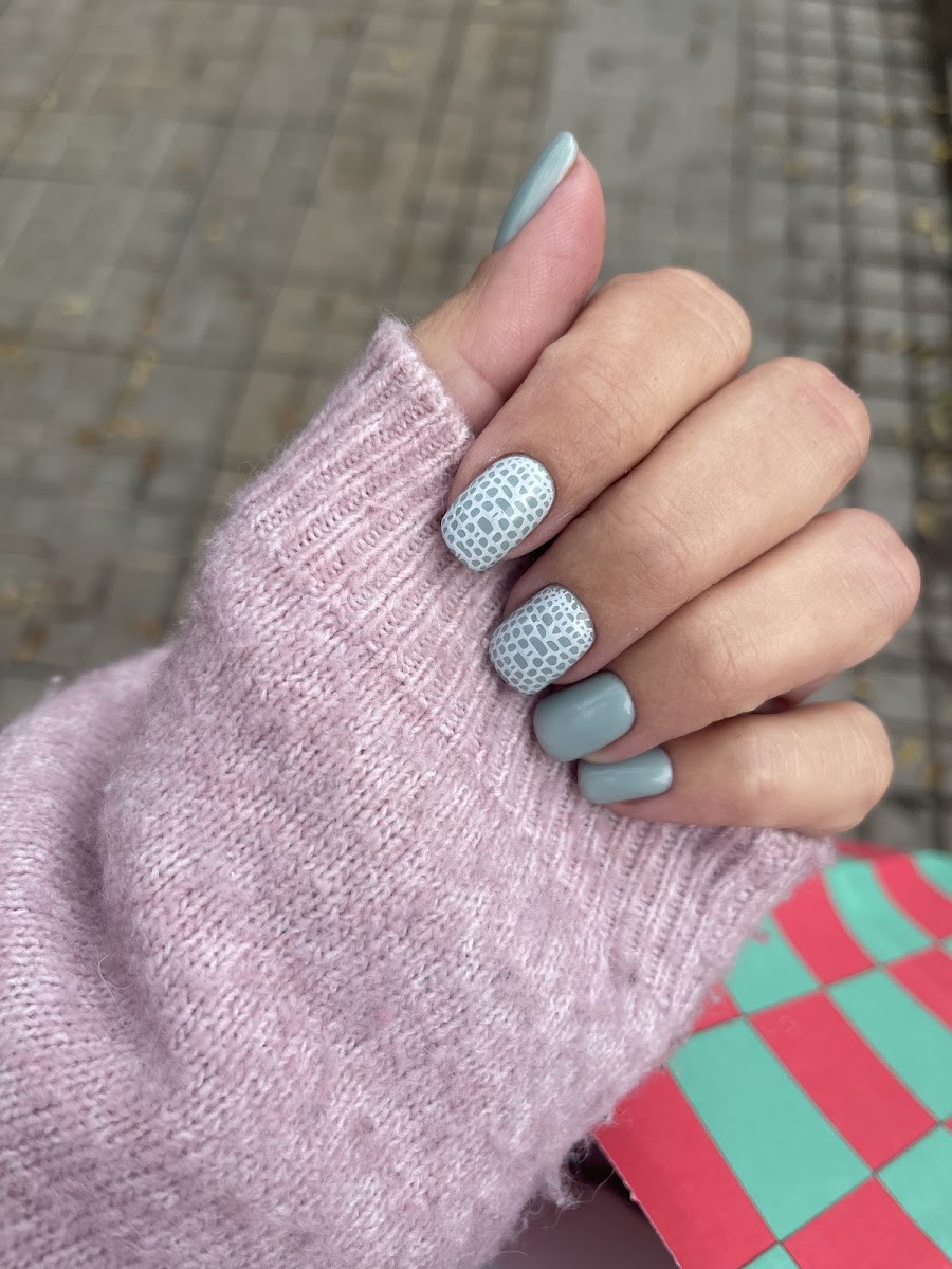 Mano con manicura en azul pastel y gris, con diseño de encaje en dos uñas, sobre un jersey rosa