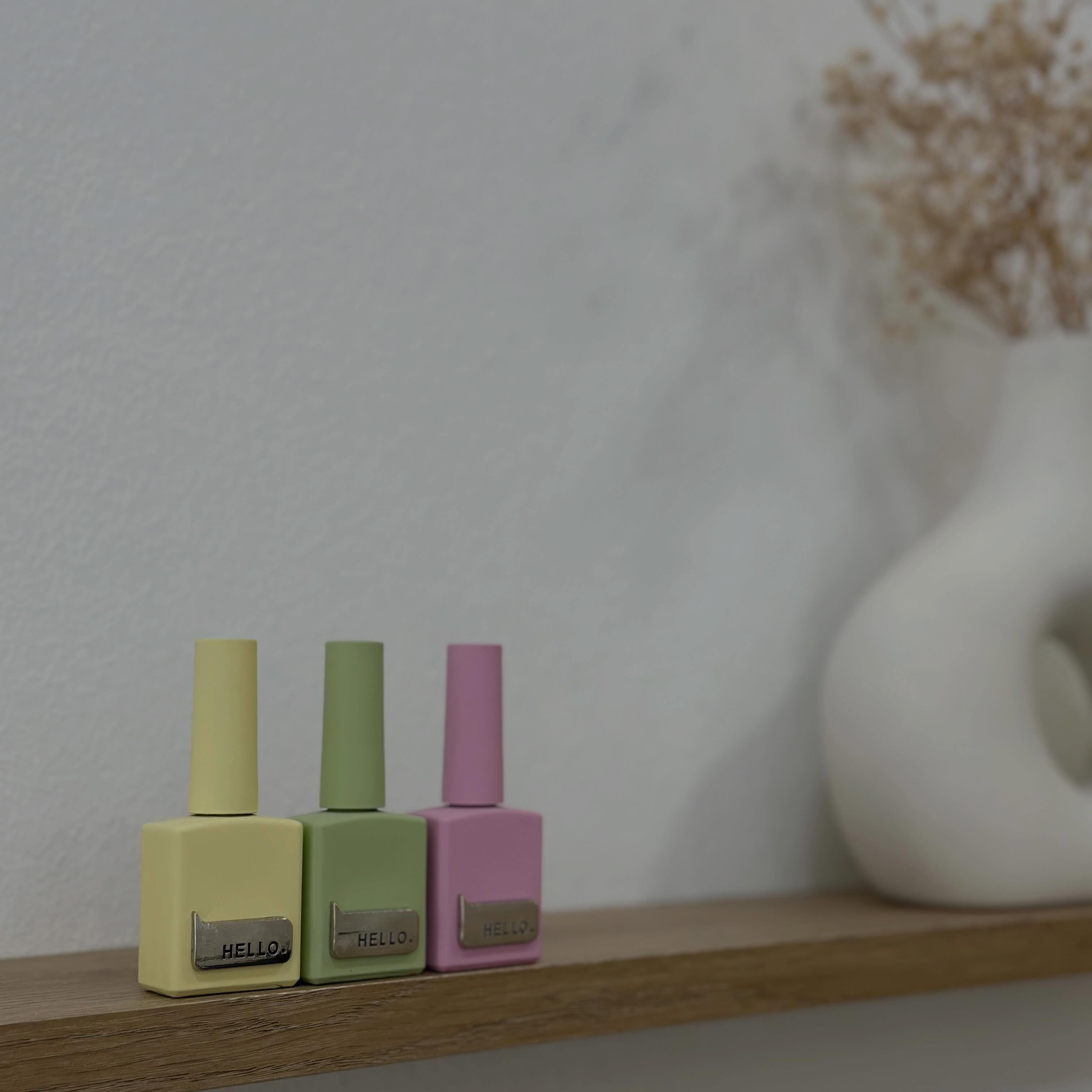 Tres botes de esmalte en amarillo, verde y rosa pastel con la etiqueta «HELLO» sobre una superficie de madera