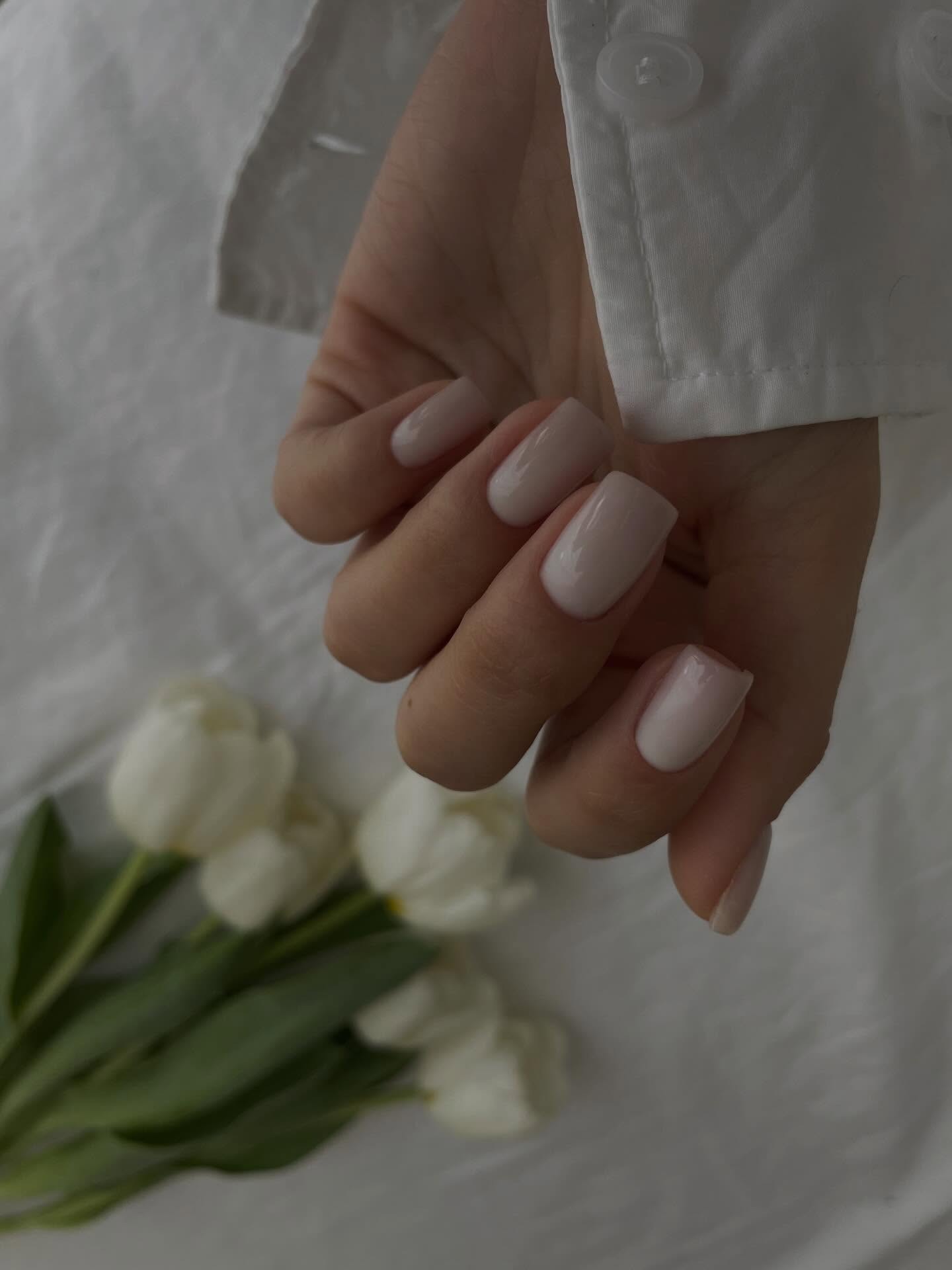 Mano con manicura corta en blanco rosado sobre una camisa blanca, con un ramo de flores blancas al fondo