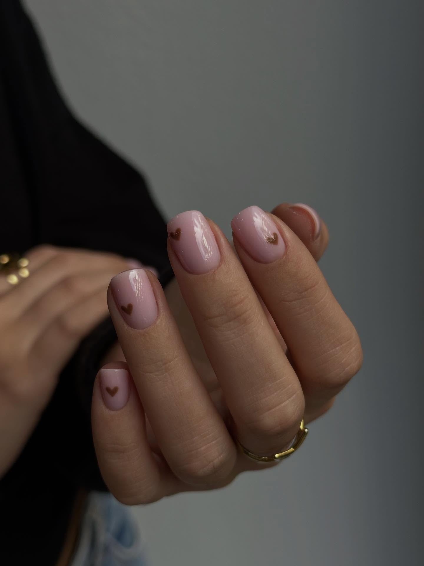 Mano con manicura rosa palo y pequeños corazones marrones, anillos dorados y fondo gris