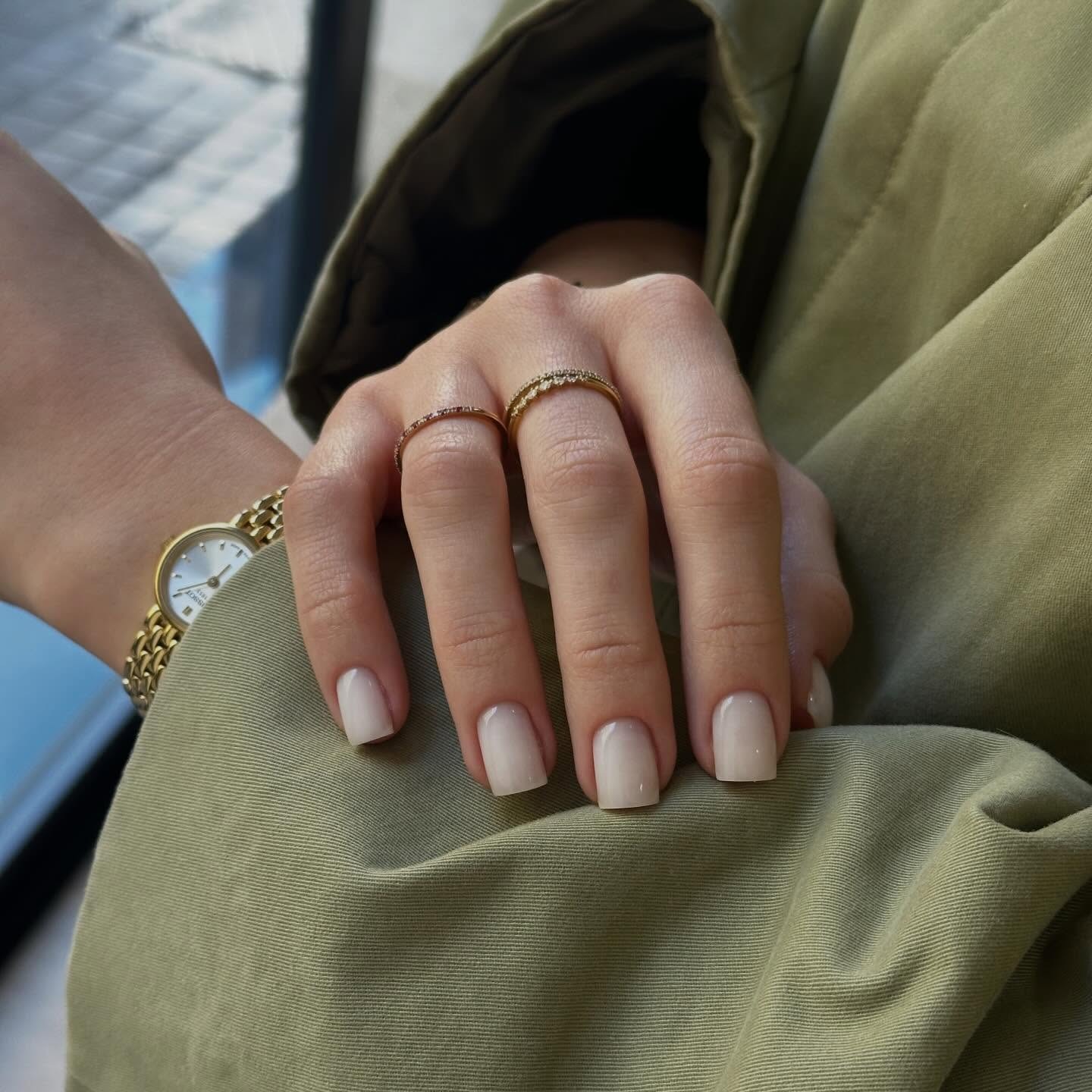 Mano con manicura nude y dos anillos dorados sobre una prenda verde oliva, con reloj dorado en la muñeca