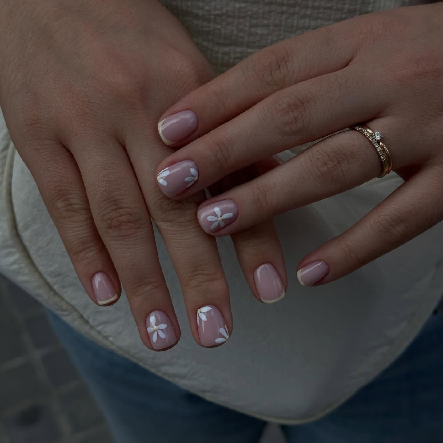 Manos juntas con manicura corta y flores blancas pintadas, anillos dorados con piedras sobre una superficie beige