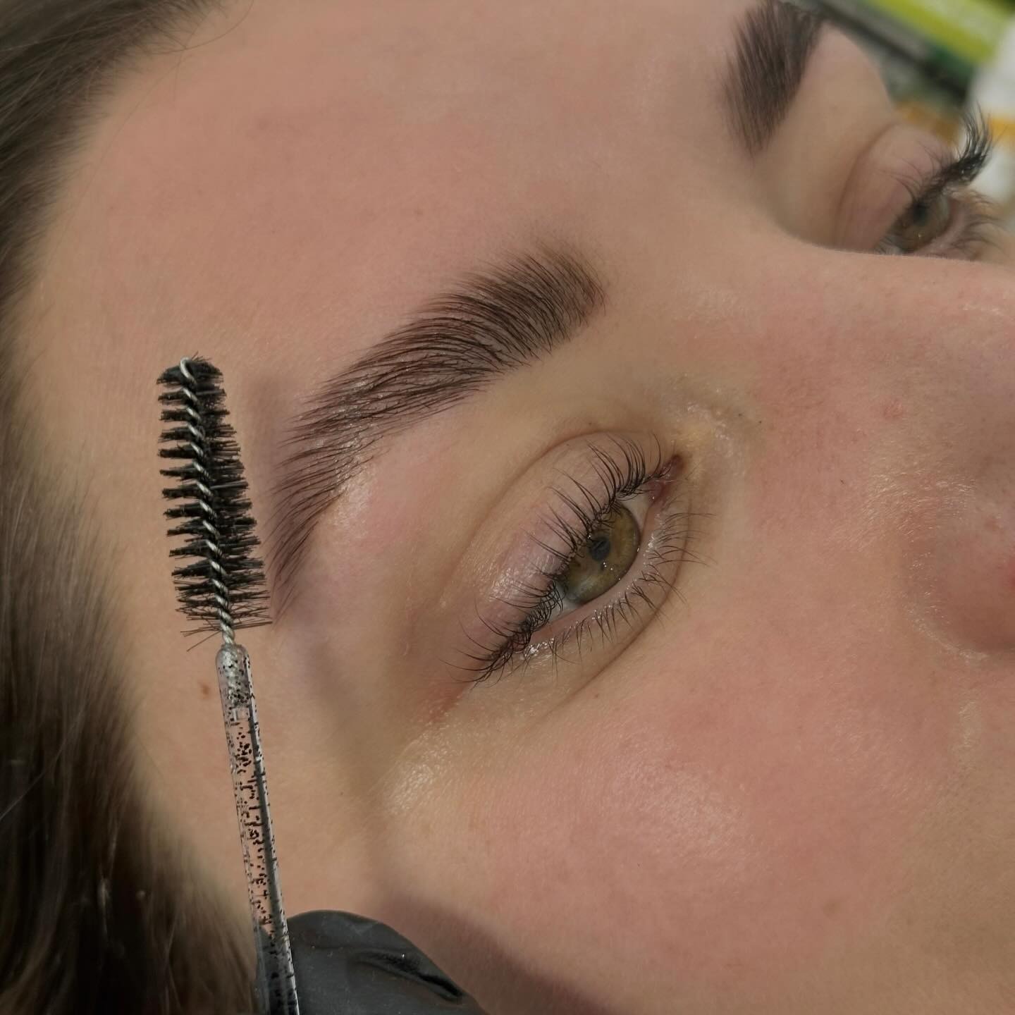 Primer plano de un rostro con las cejas peinadas hacia arriba, extensiones de pestañas y una brocha de rímel