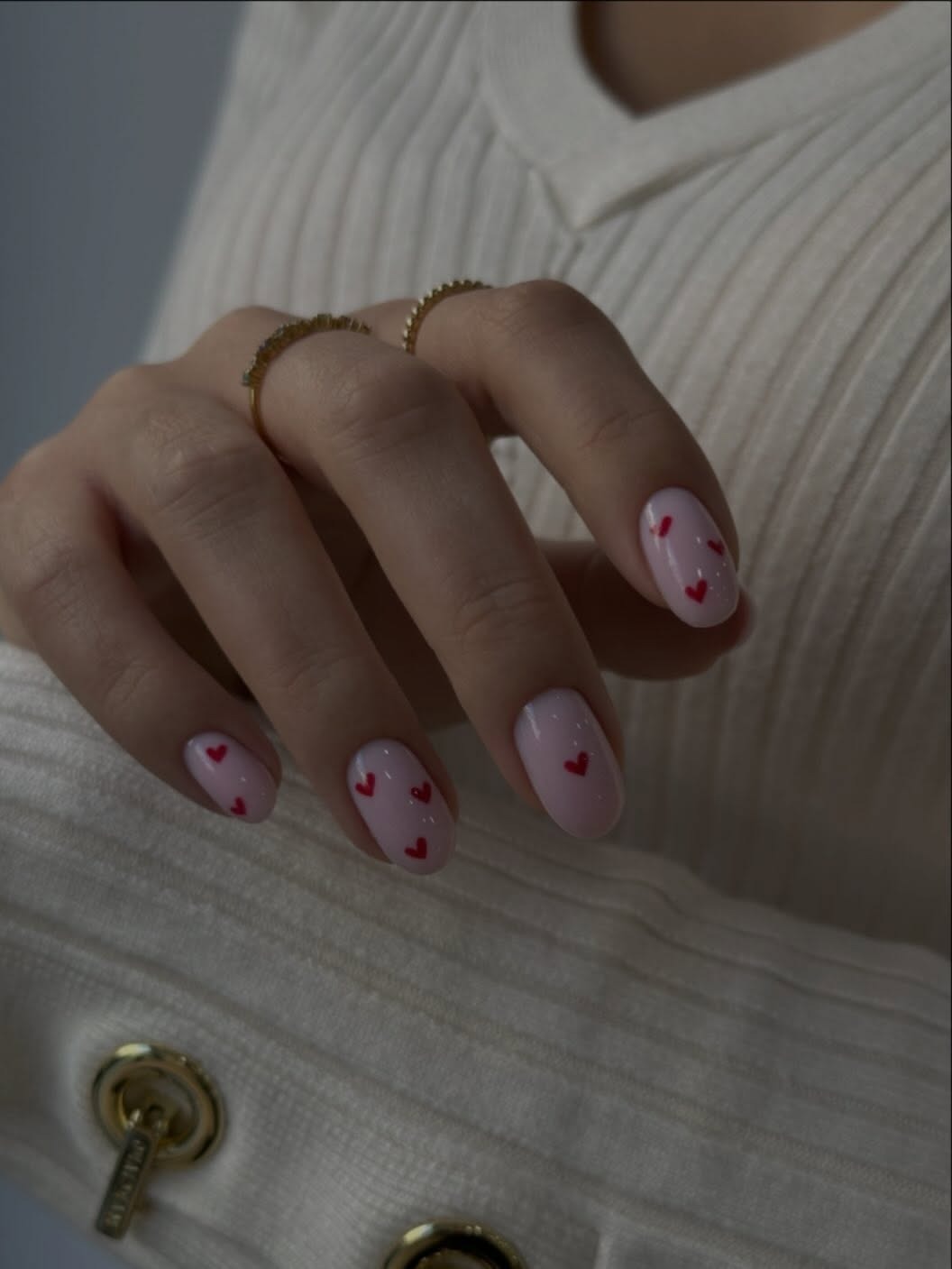 Mano con manicura rosa claro decorada con pequeños corazones rojos y dos anillos dorados sobre un jersey blanco de punto